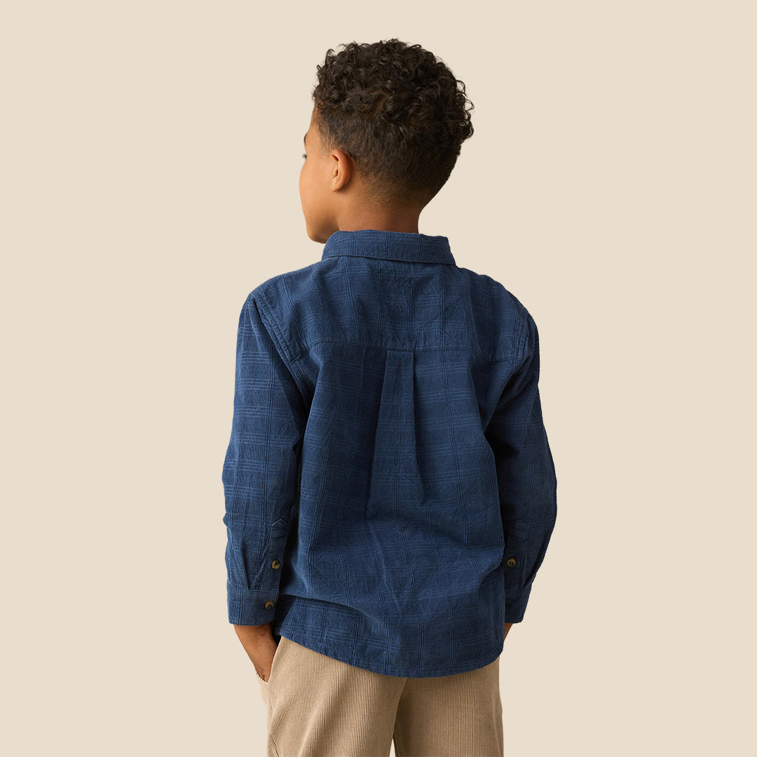 A young boy with curly dark hair wearing a dark blue long-sleeve button-up shirt and khaki pants, standing against a solid beige background.