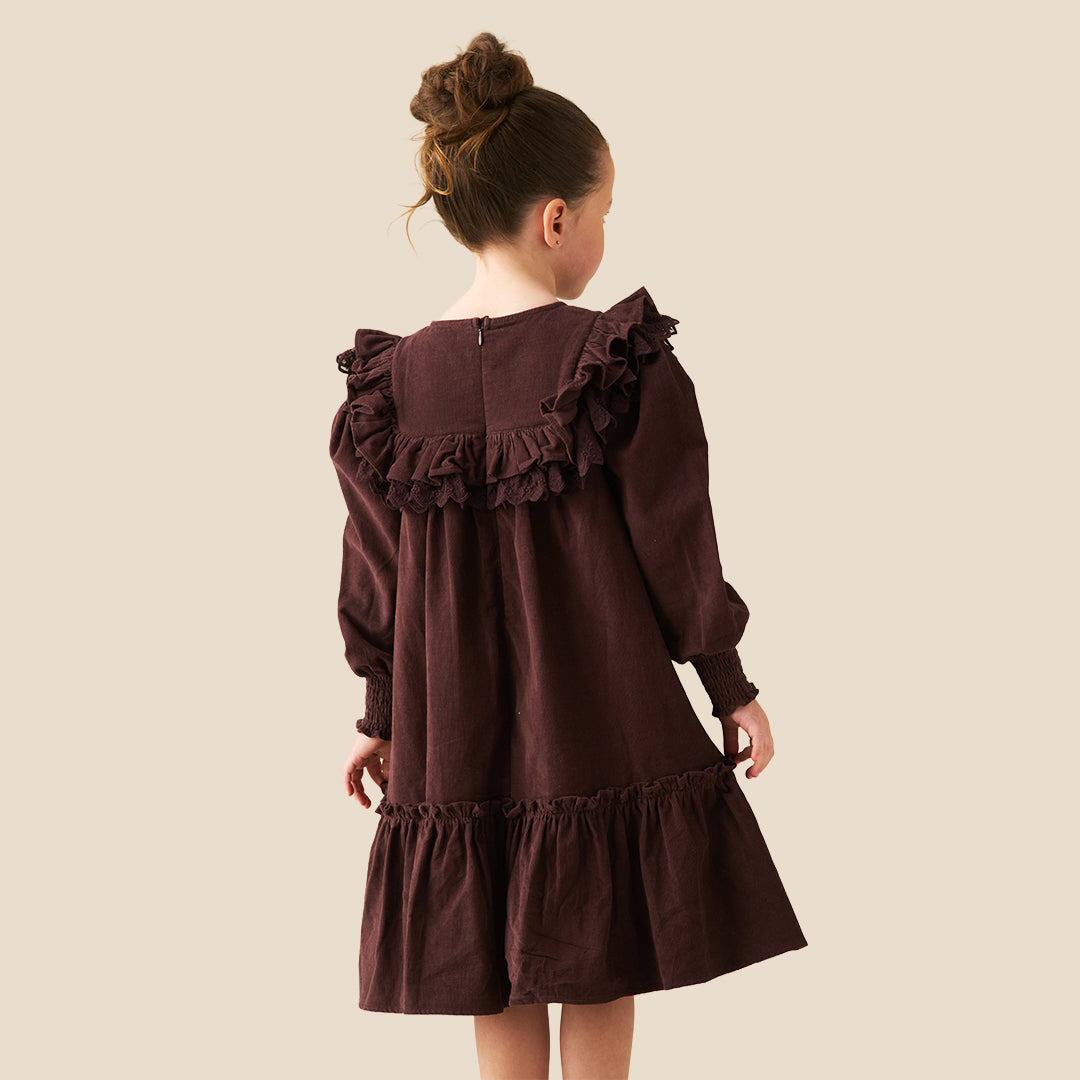 A young girl with her hair in a high bun models a dark brown long-sleeve dress with ruffled details, set against a solid light beige background.