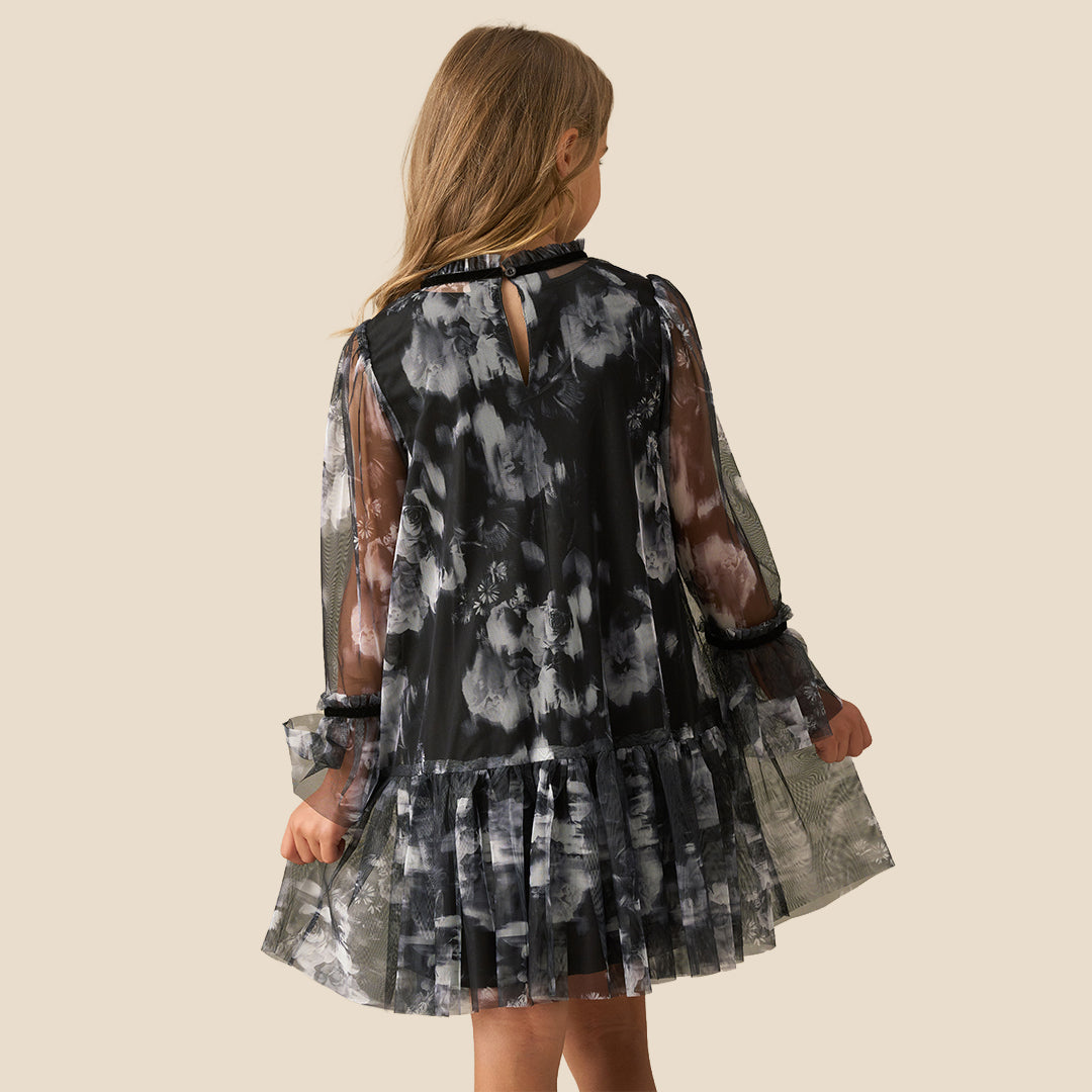 A young girl with long light brown hair is wearing a black and gray tie-dye dress with sheer long sleeves and a ruffled skirt, standing against a solid beige background.