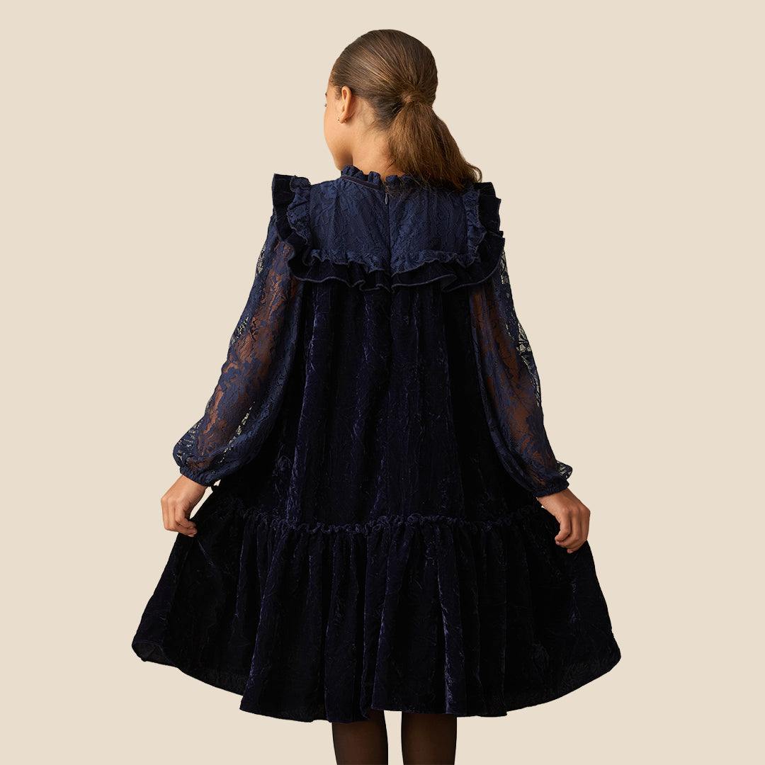 A young girl with her hair in a ponytail models a dark navy blue dress. The dress features a velvet-like texture, a ruffled collar, sheer lace long sleeves, and a tiered skirt, set against a solid beige background.