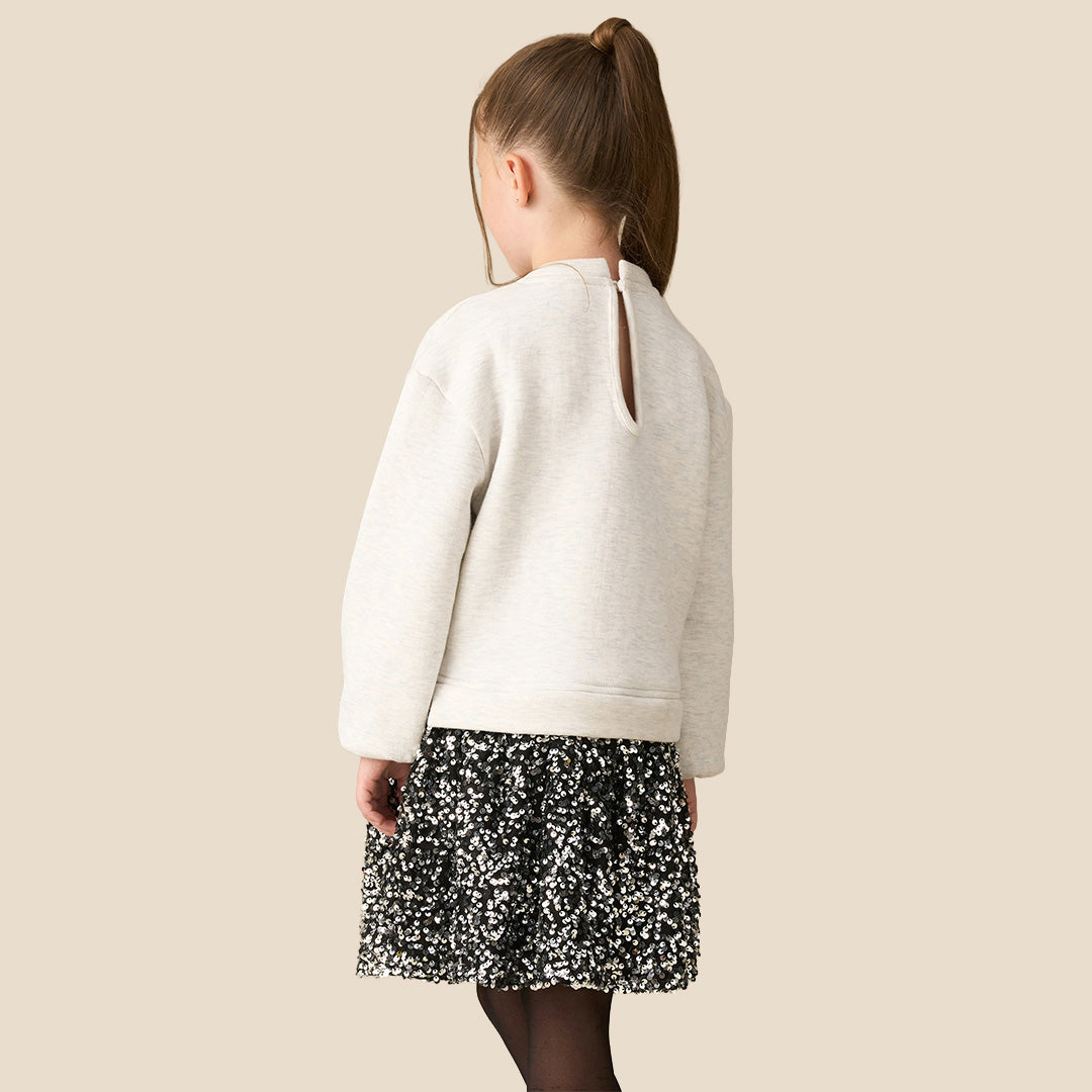 A young girl modeling a cream-colored knit sweater with a large matching fabric bow at the waist, paired with a black and white floral patterned skirt, against a solid beige background.