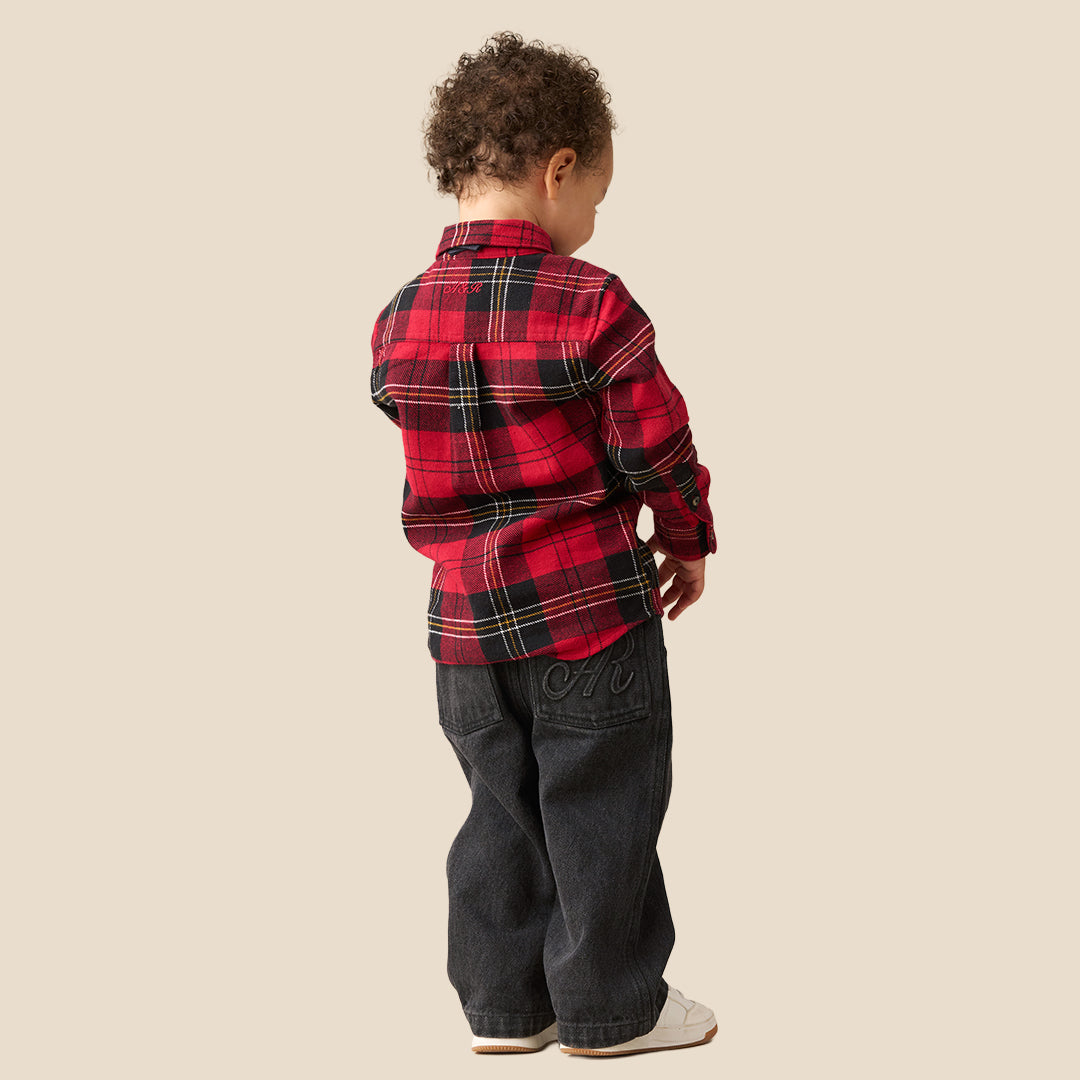 A toddler standing against a plain beige background, wearing a red and black plaid long-sleeve shirt with a matching dark bow tie, dark pants, and white sneakers.