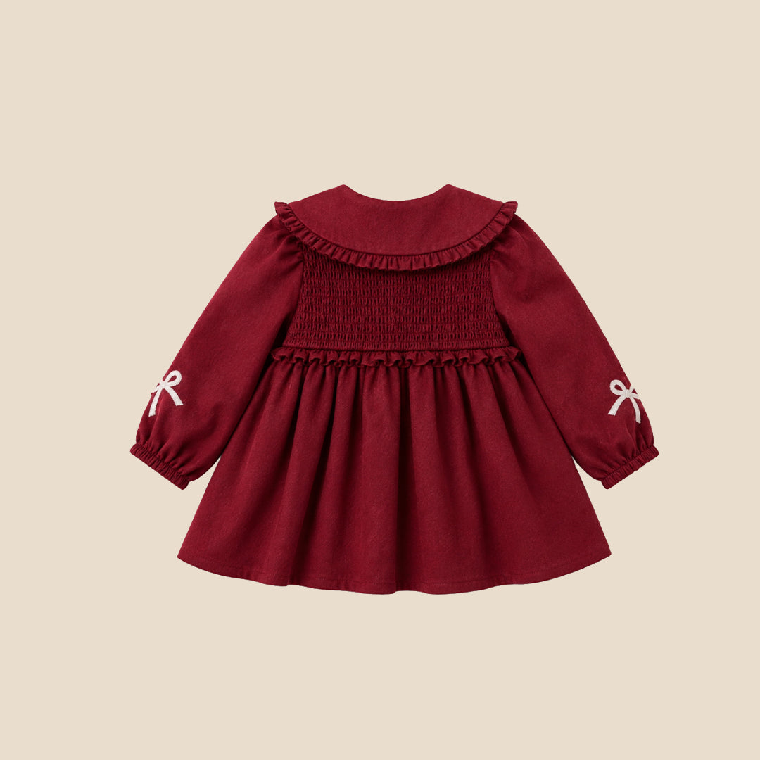 A toddler girl wearing a burgundy corduroy dress with a large white ruffled collar decorated with bows, white tights, and beige ballet flats, standing against a solid beige background.