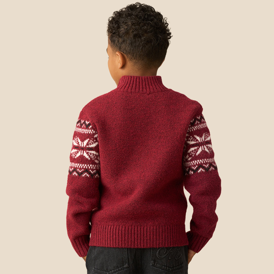 A young boy smiling and wearing a burgundy knit quarter-zip sweater with a white and dark red Fair Isle snowflake pattern across the chest and shoulders, set against a solid beige background.