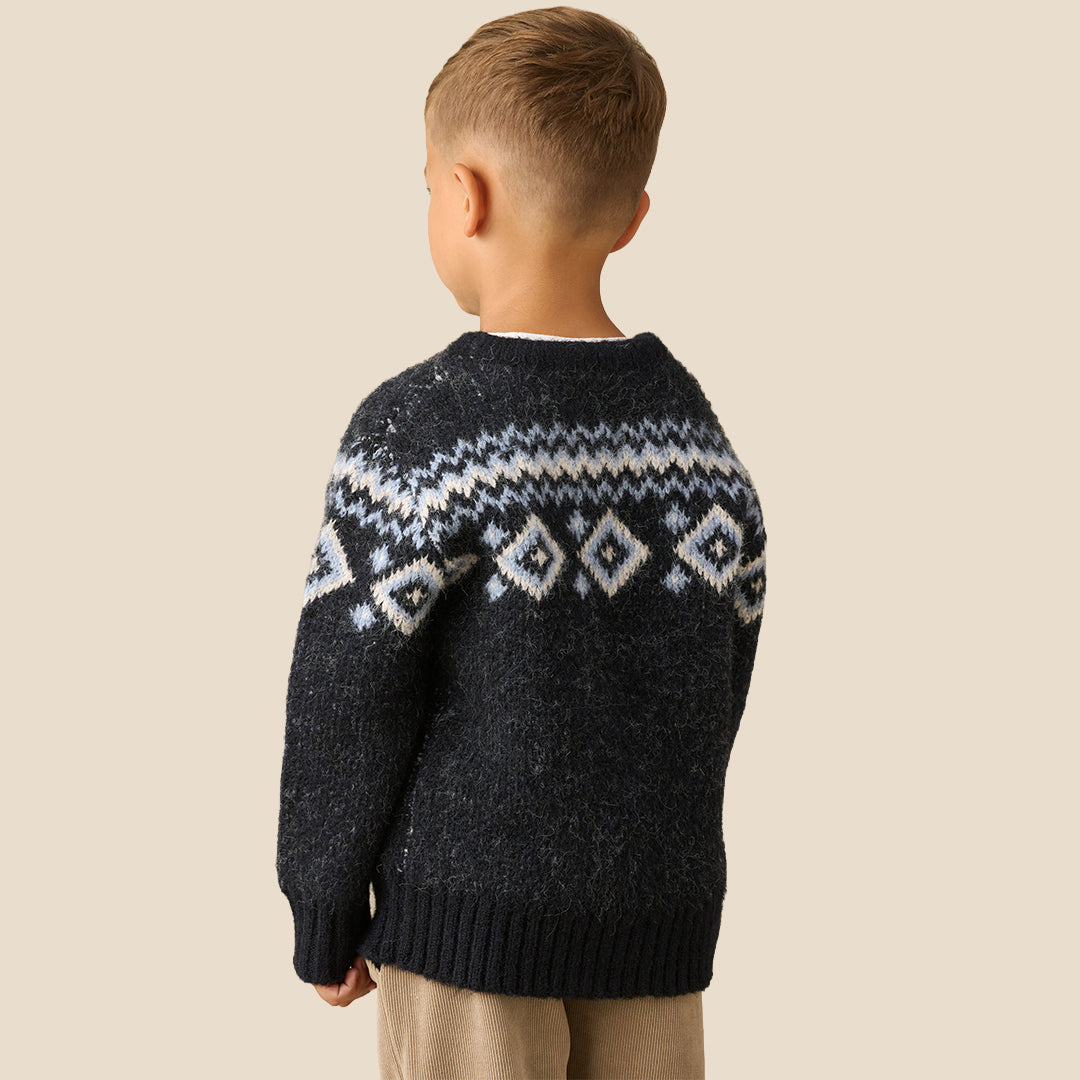 A young boy wearing a dark navy knit sweater with a white and light blue geometric Fair Isle pattern across the chest and shoulders, paired with beige pants, set against a solid light beige background.