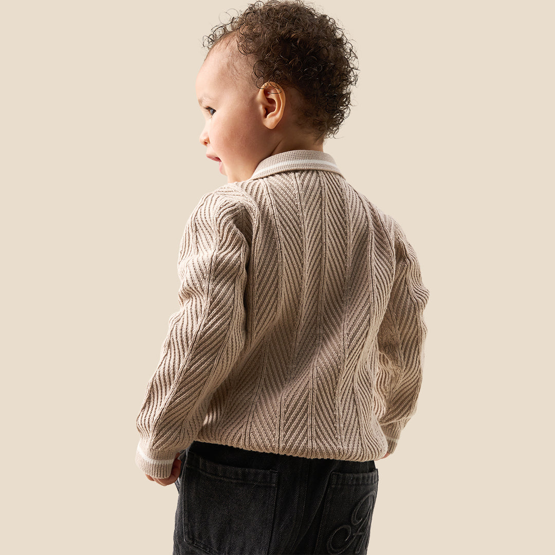 A toddler with curly brown hair wearing a cream-colored cable-knit sweater with a white Peter Pan collar, standing against a solid beige background.