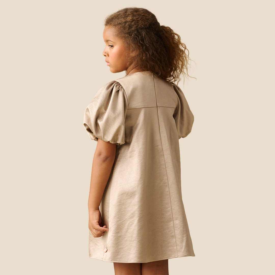 A young girl with curly brown hair wearing a champagne beige dress with puffed short sleeves and a large bow at the neckline, standing against a solid light beige background.