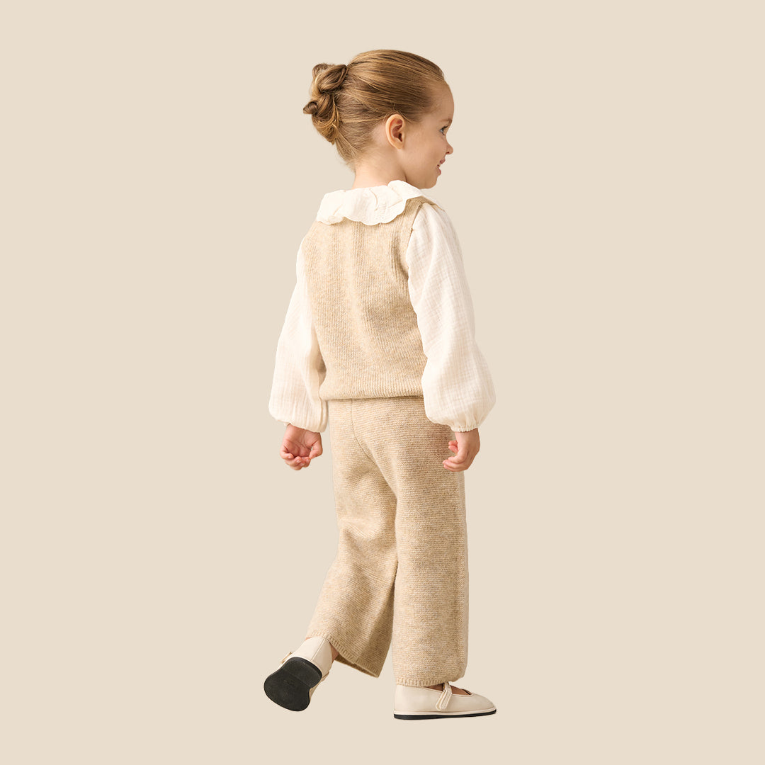 A young child modeling a cream and beige knit outfit, consisting of a long-sleeve top with a ruffled collar and matching knit vest and pants, set against a solid light beige background.