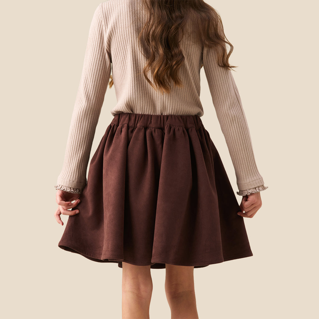 A model wearing a beige ribbed cardigan and a dark brown pleated skirt with a large bow at the waist, shown from the chest down against a solid beige background.