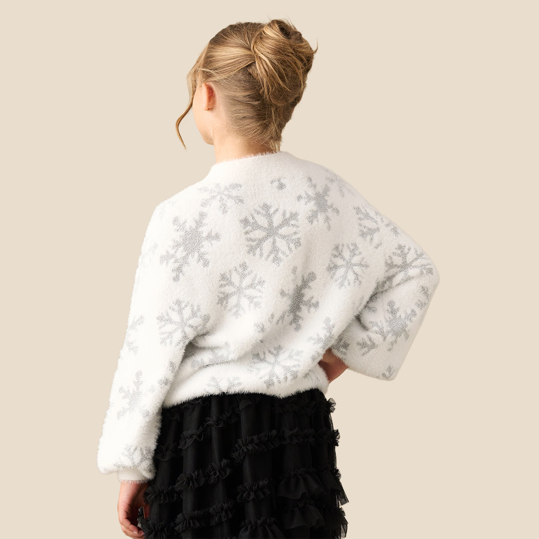 A young girl with blonde hair in a bun wearing a fuzzy white long-sleeve sweater with a gray snowflake pattern, paired with a black ruffled skirt, set against a solid beige background.