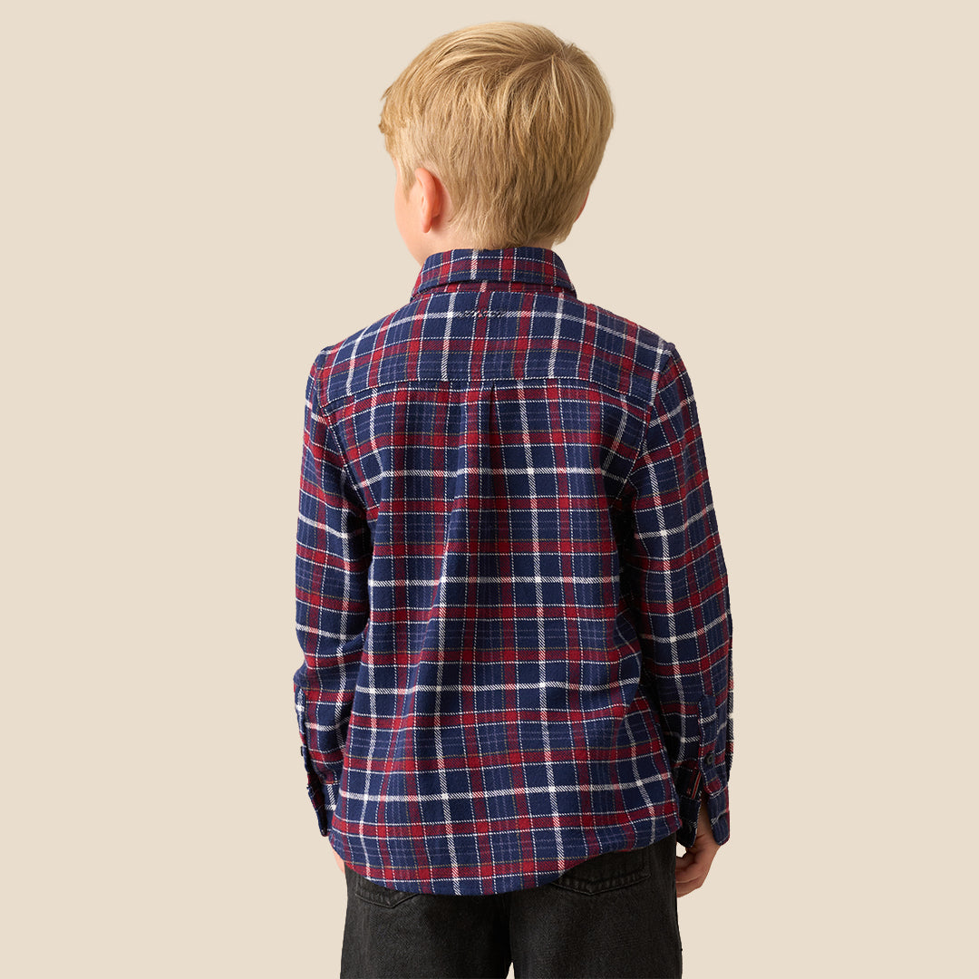 A young boy with blond hair wearing a red, navy, and white plaid long-sleeve button-up shirt with a black bow tie, paired with black pants, standing against a solid beige background.