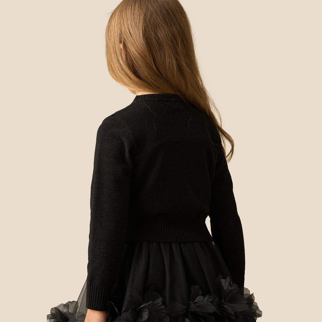 A young girl with long light brown hair wearing a black knit cardigan with gold bow-shaped buttons and a black tulle skirt, standing against a solid beige background.