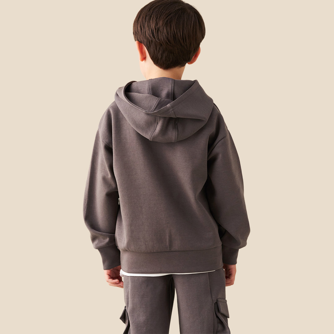 A young boy wearing a dark gray hoodie with a small white graphic on the chest and matching dark gray cargo pants, standing against a solid beige background.