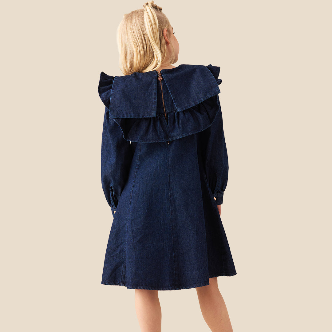 A young girl with blonde hair in two buns wearing a dark blue denim dress with a large ruffled collar, standing against a solid beige background.