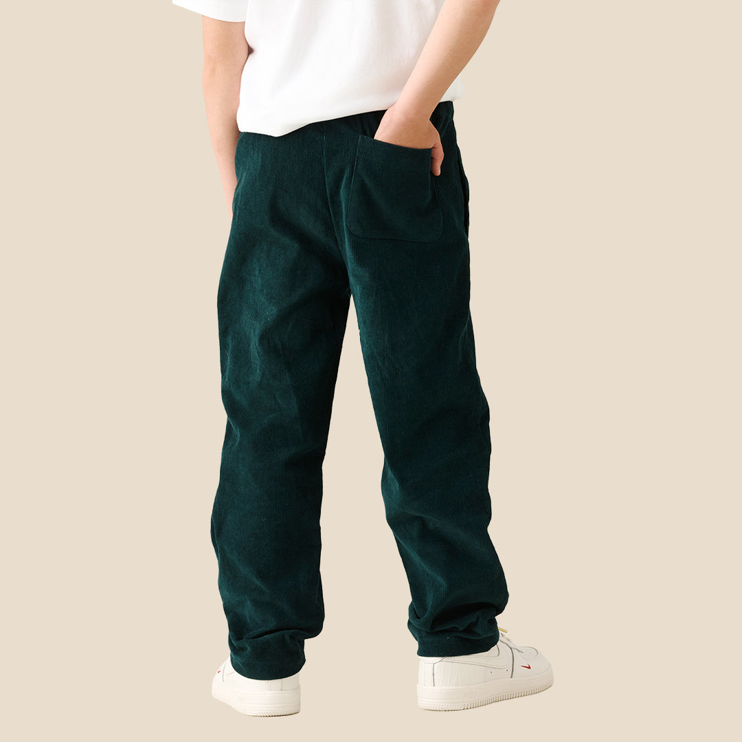 A young boy modeling a white crewneck t-shirt and dark green corduroy pants, standing against a solid beige background.