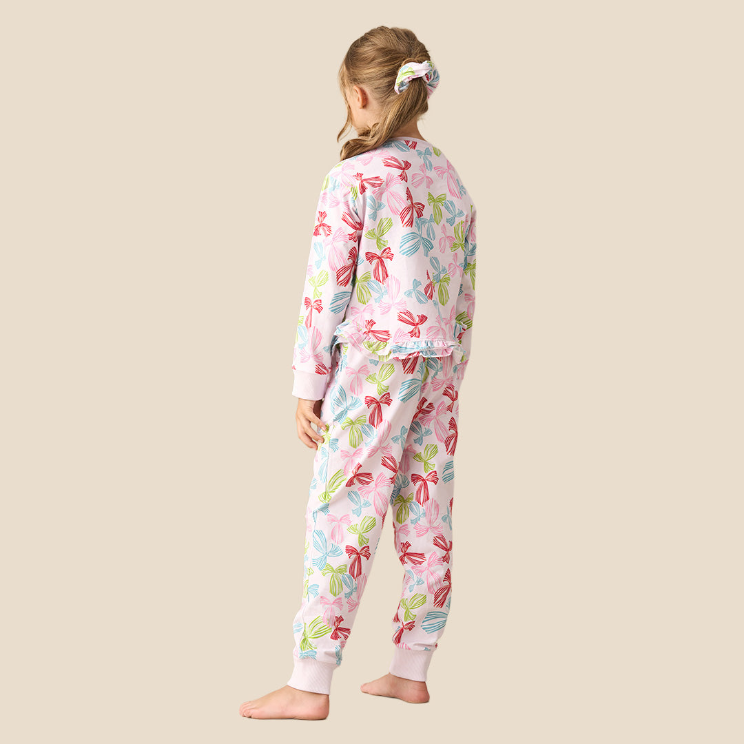 A young girl modeling a two-piece pajama set featuring a long-sleeve top and matching pants with a colorful bow pattern on a light pink background, set against a solid beige backdrop.