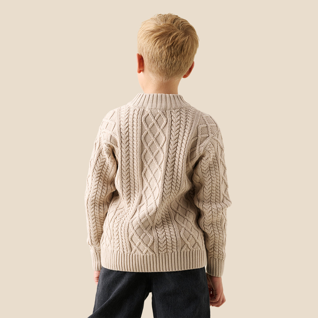 A young boy wearing a beige cable-knit zip-up cardigan over a white t-shirt and dark jeans, standing against a solid light beige background.