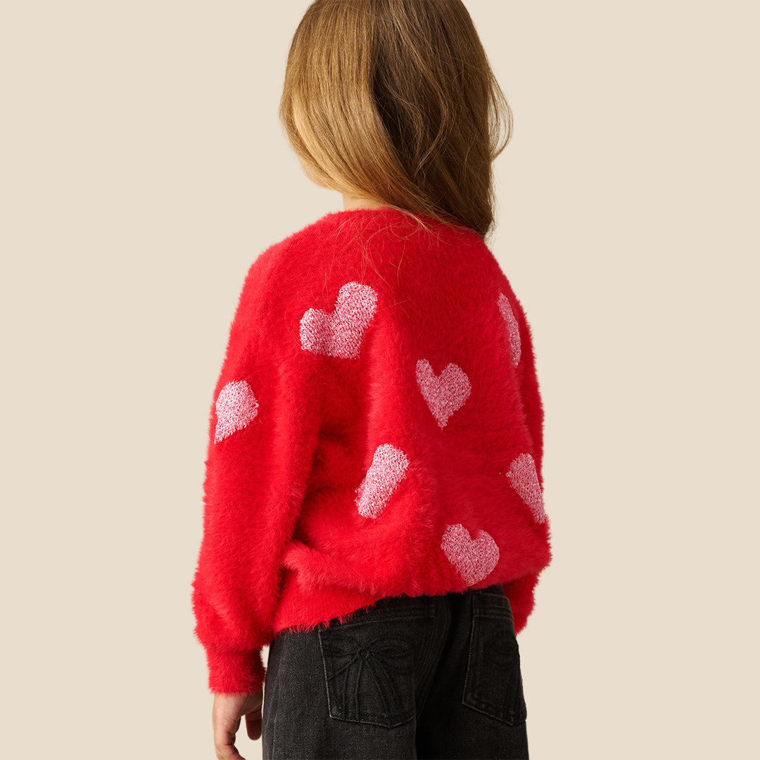 A young girl with long light brown hair wearing a fuzzy red knit sweater with light pink hearts, posing against a solid beige background.