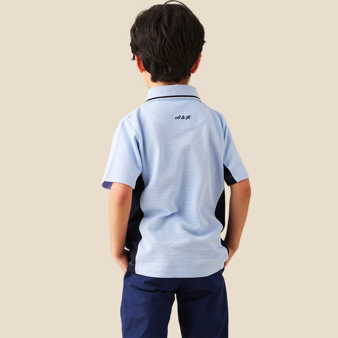 A young boy modeling a light blue polo shirt with dark navy side panels and a half-zip collar, paired with navy pants, against a solid beige background.
