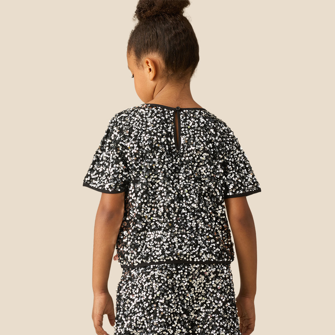 A young girl with her hair in a high bun is wearing a short-sleeve black dress covered in a dense white floral pattern, posed against a solid beige background.