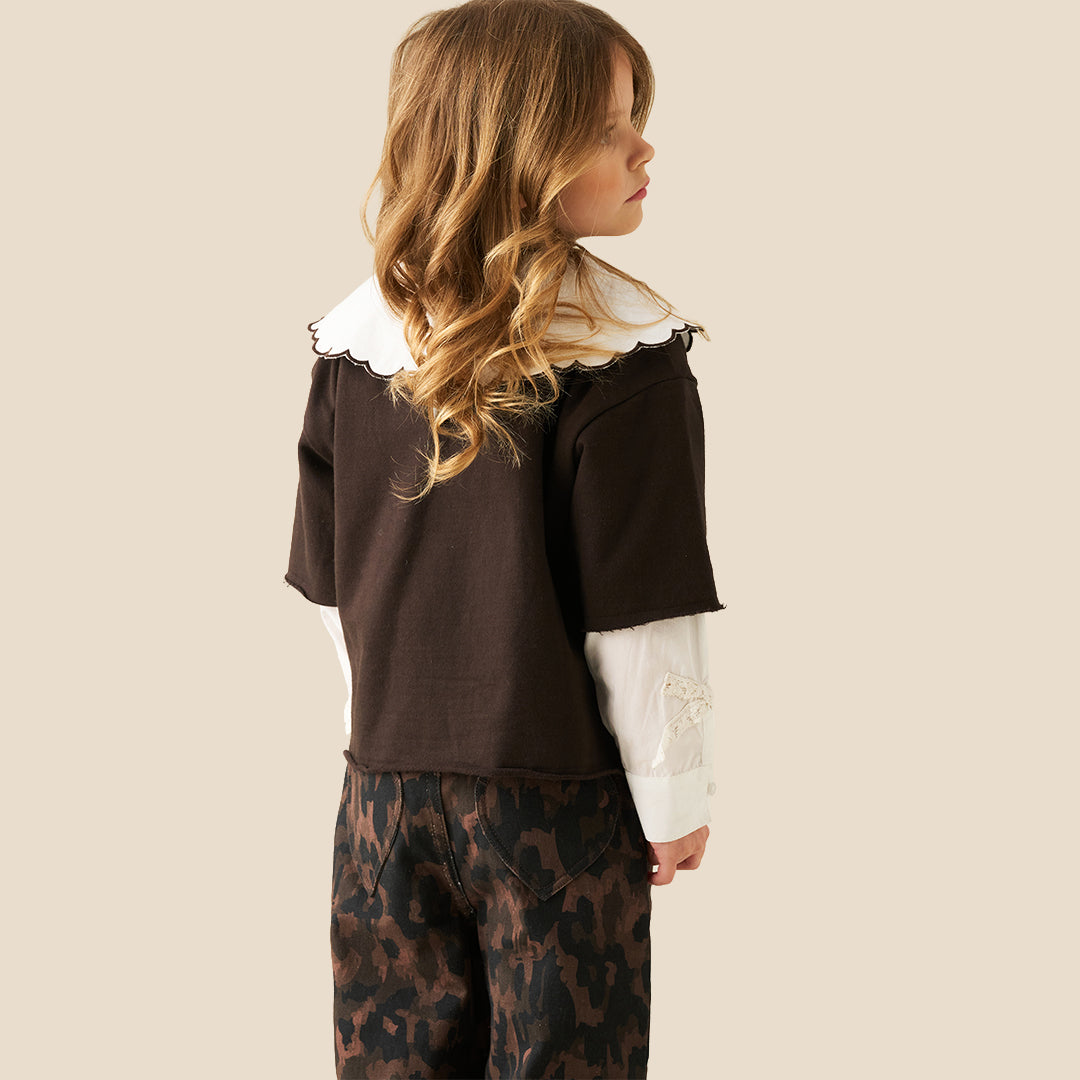 A young girl with wavy light brown hair is wearing a dark brown short-sleeve top with a large white ruffled collar featuring black cursive embroidery, layered over a white long-sleeve shirt, paired with brown and black leopard-print pants, against a solid beige background.