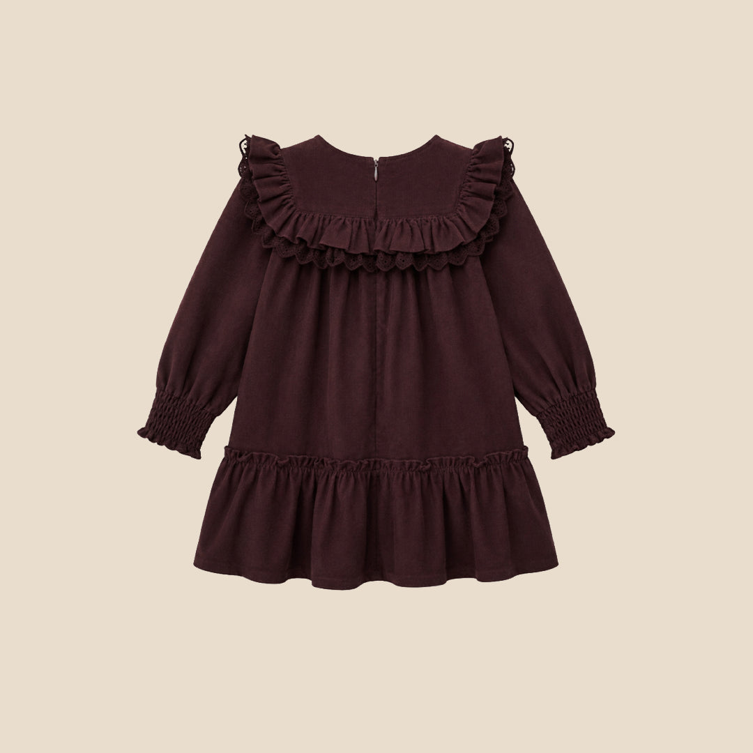 A young girl with light brown hair in pigtails stands against a solid beige background wearing a long-sleeved dark plum-brown dress with ruffles, white tights, and cream-colored shoes.