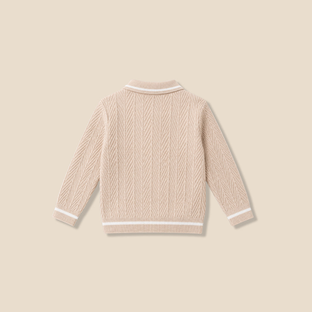 A toddler with curly brown hair wearing a cream-colored cable-knit sweater with a white Peter Pan collar, standing against a solid beige background.