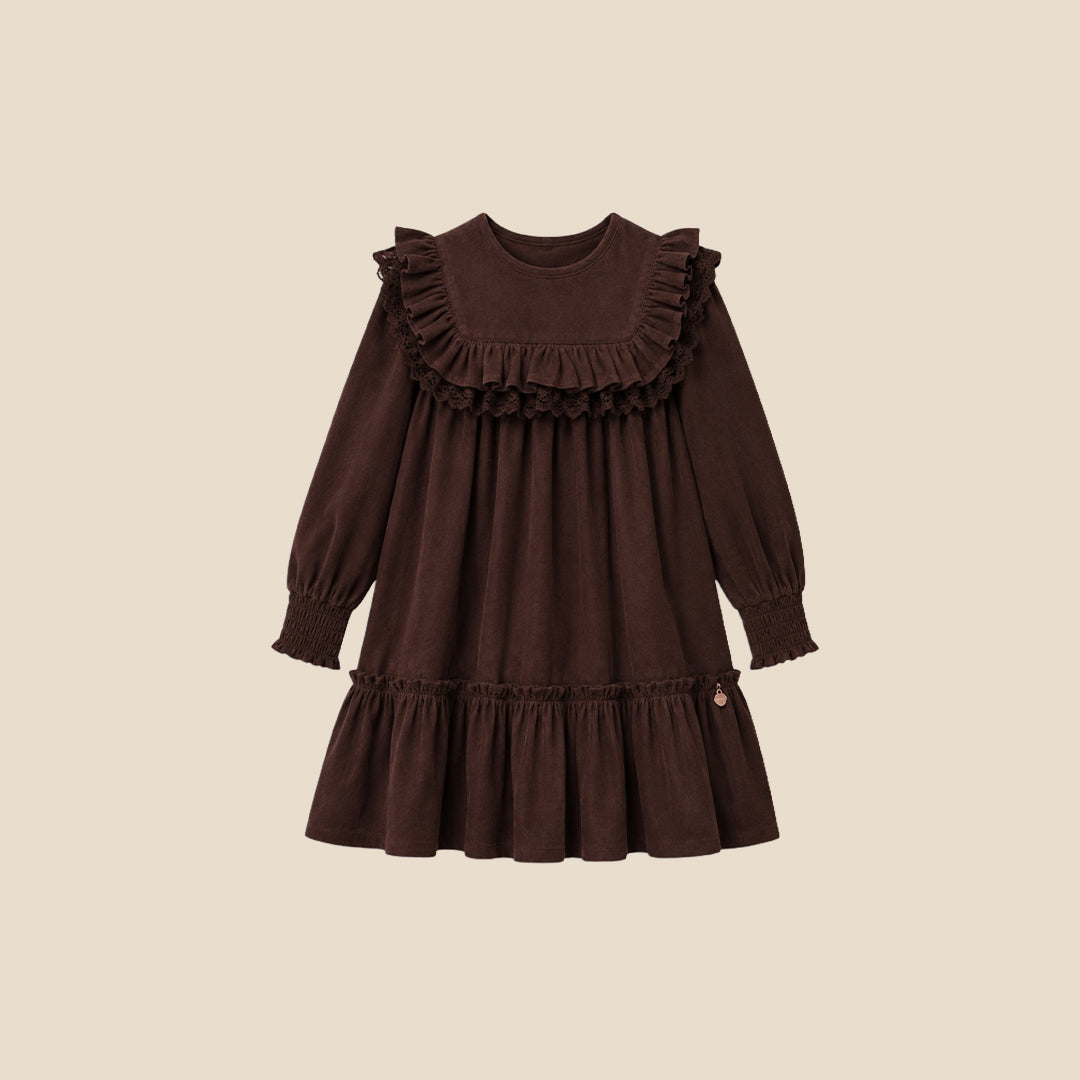 A young girl with her hair in a high bun models a dark brown long-sleeve dress with ruffled details, set against a solid light beige background.