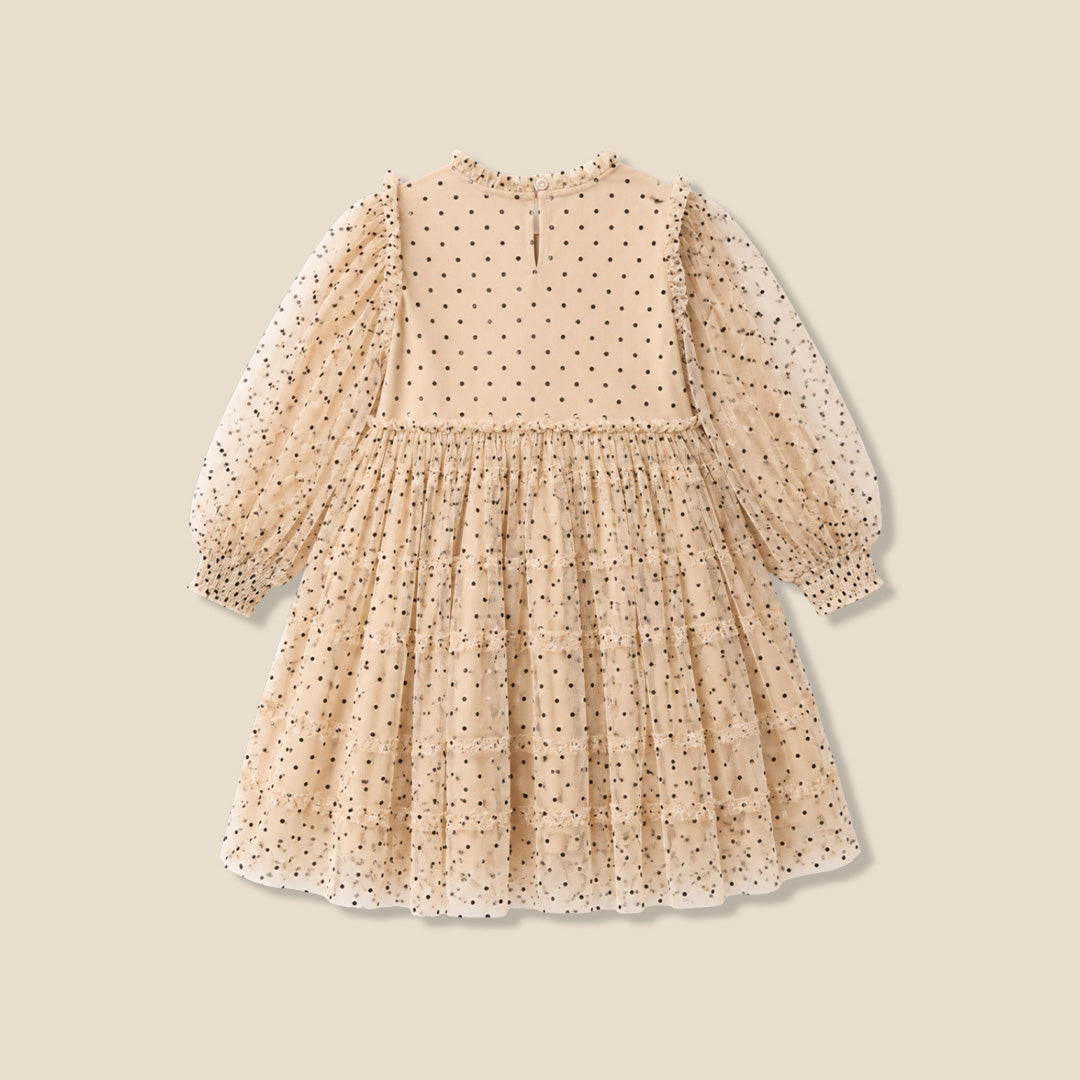 A young girl with her hair in a bun wears a cream-colored, long-sleeved tiered dress with black polka dots, standing against a solid beige background.