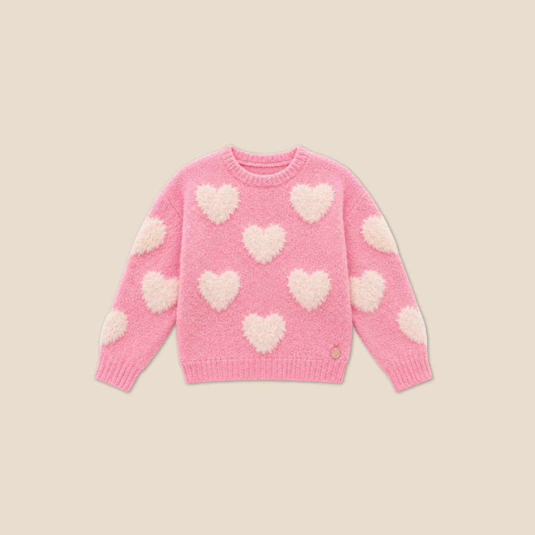 A young girl with blonde hair styled in buns wears a pink fuzzy knit sweater with fluffy white heart details, posing with her hands forming a heart shape in front of her chest. She is wearing a dark blue denim skirt.
