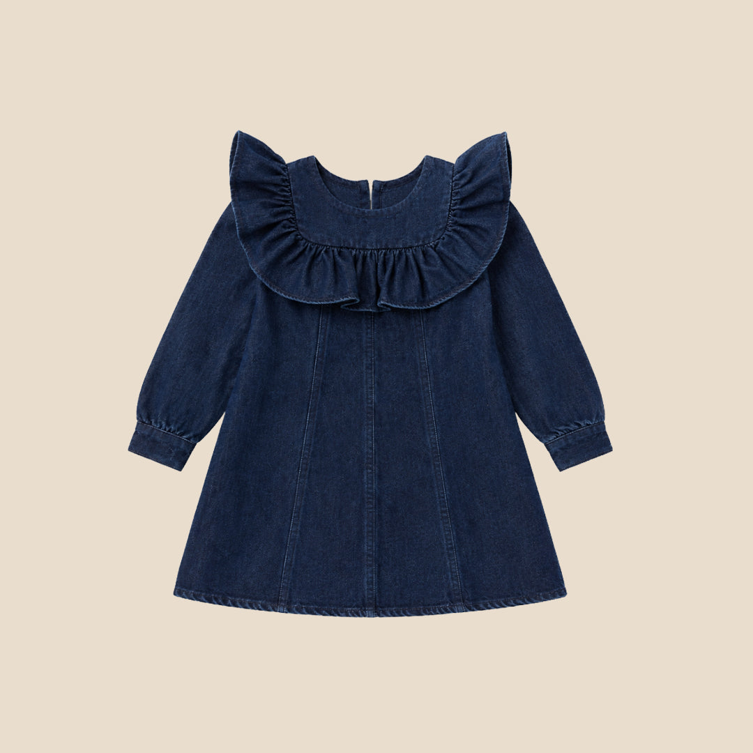 A young child wearing a dark blue denim dress with dramatic ruffled shoulders, standing against a solid beige background.