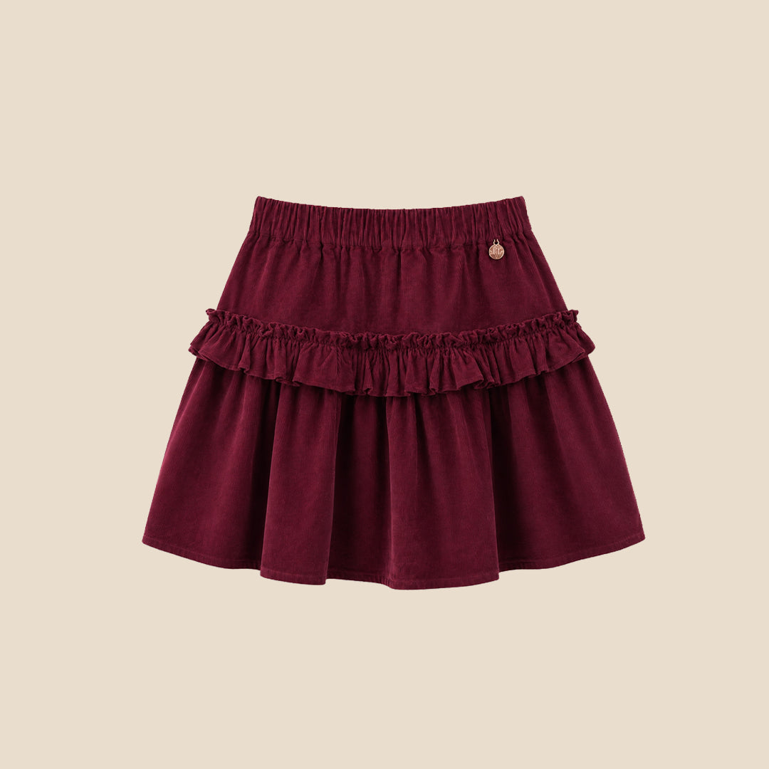 A child wearing a tiered burgundy skirt with ruffles and a white puff-sleeve top featuring a pink plaid bow graphic, photographed against a plain beige background.