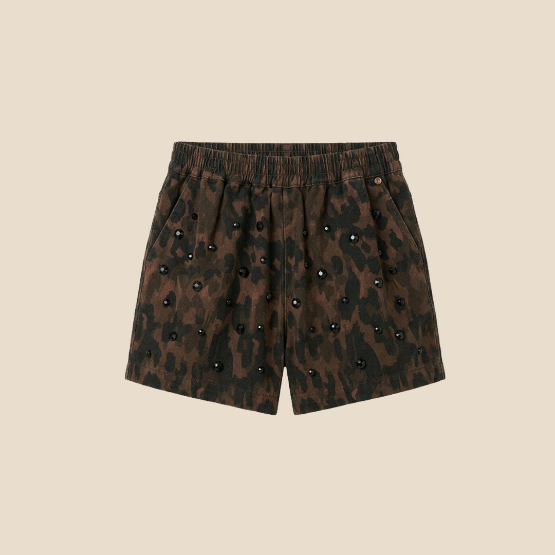 A child wearing a matching short-sleeve button-front top and shorts set in a dark brown and black leopard print, with decorative pearl-like embellishments, photographed against a plain beige background.