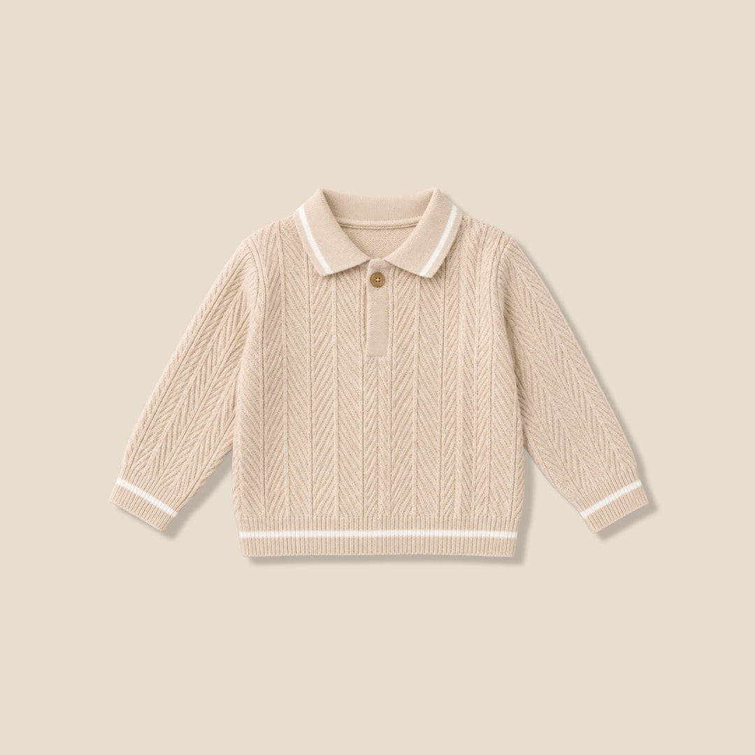 A toddler with curly brown hair wearing a cream-colored cable-knit sweater with a white Peter Pan collar, standing against a solid beige background.