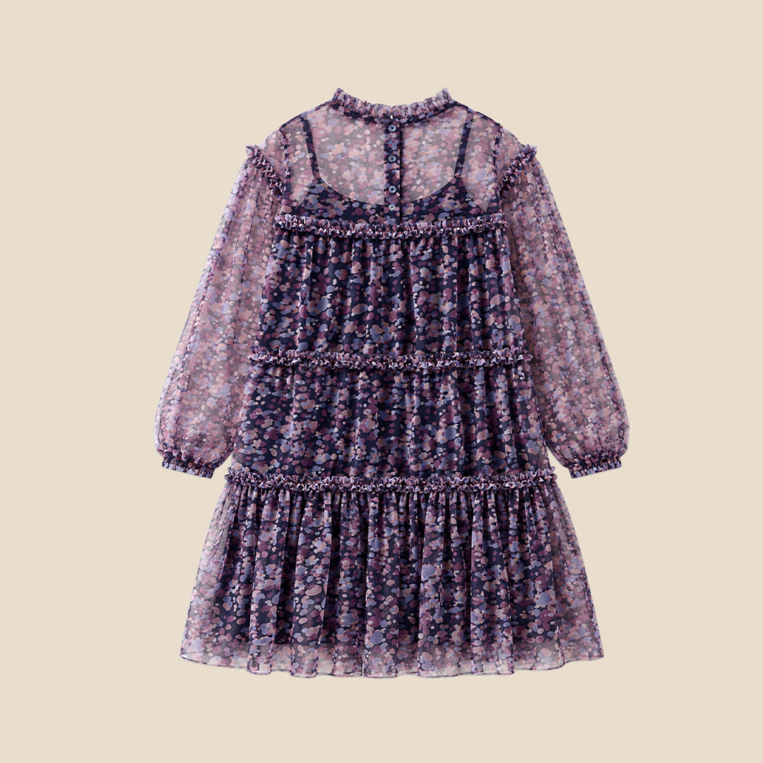 A young girl with curly hair stands on a checkered porch wearing a long-sleeved tiered dress with a pink, purple, and blue floral pattern. She is posing behind a black wrought-iron fence.
