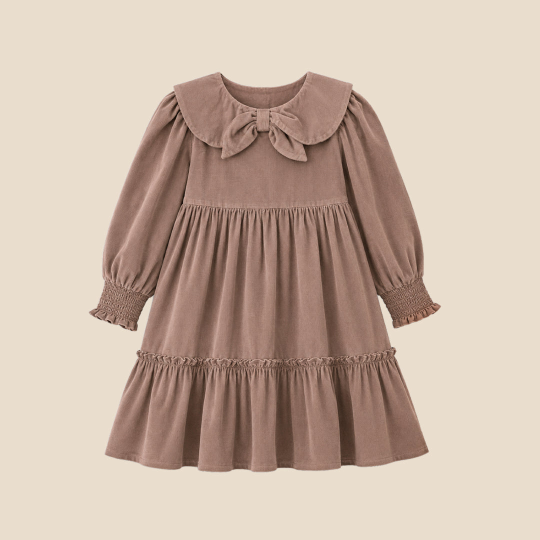 A young girl with her hair in a bun wears a brown corduroy dress with a large ruffled collar and bow, standing against a solid beige background.