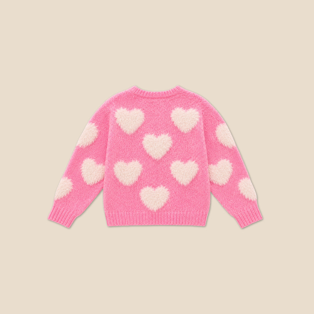 A young girl with blonde hair styled in buns wears a pink fuzzy knit sweater with fluffy white heart details, posing with her hands forming a heart shape in front of her chest. She is wearing a dark blue denim skirt.