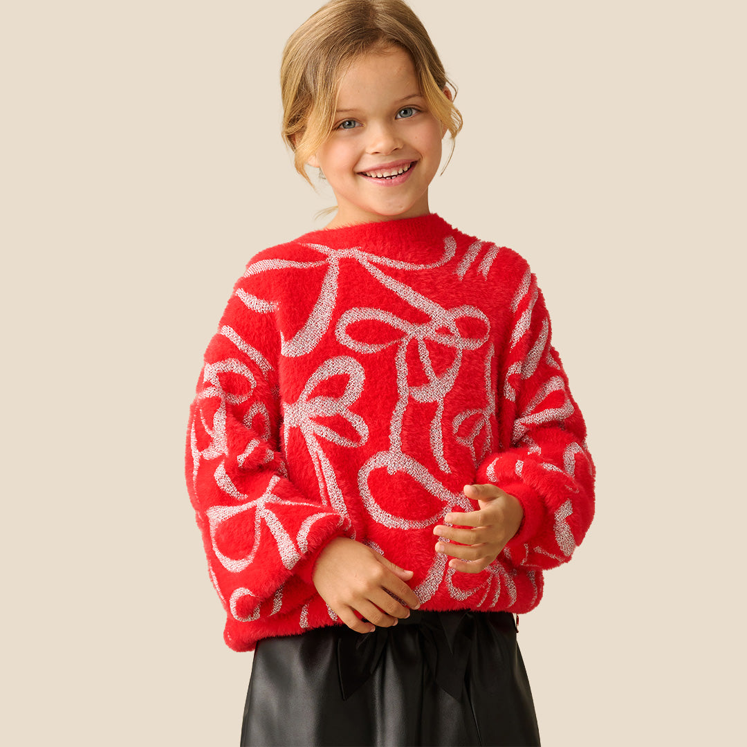A young girl smiling while wearing a bright red fuzzy knit sweater with a white abstract pattern, paired with black bottoms, against a solid beige background.
