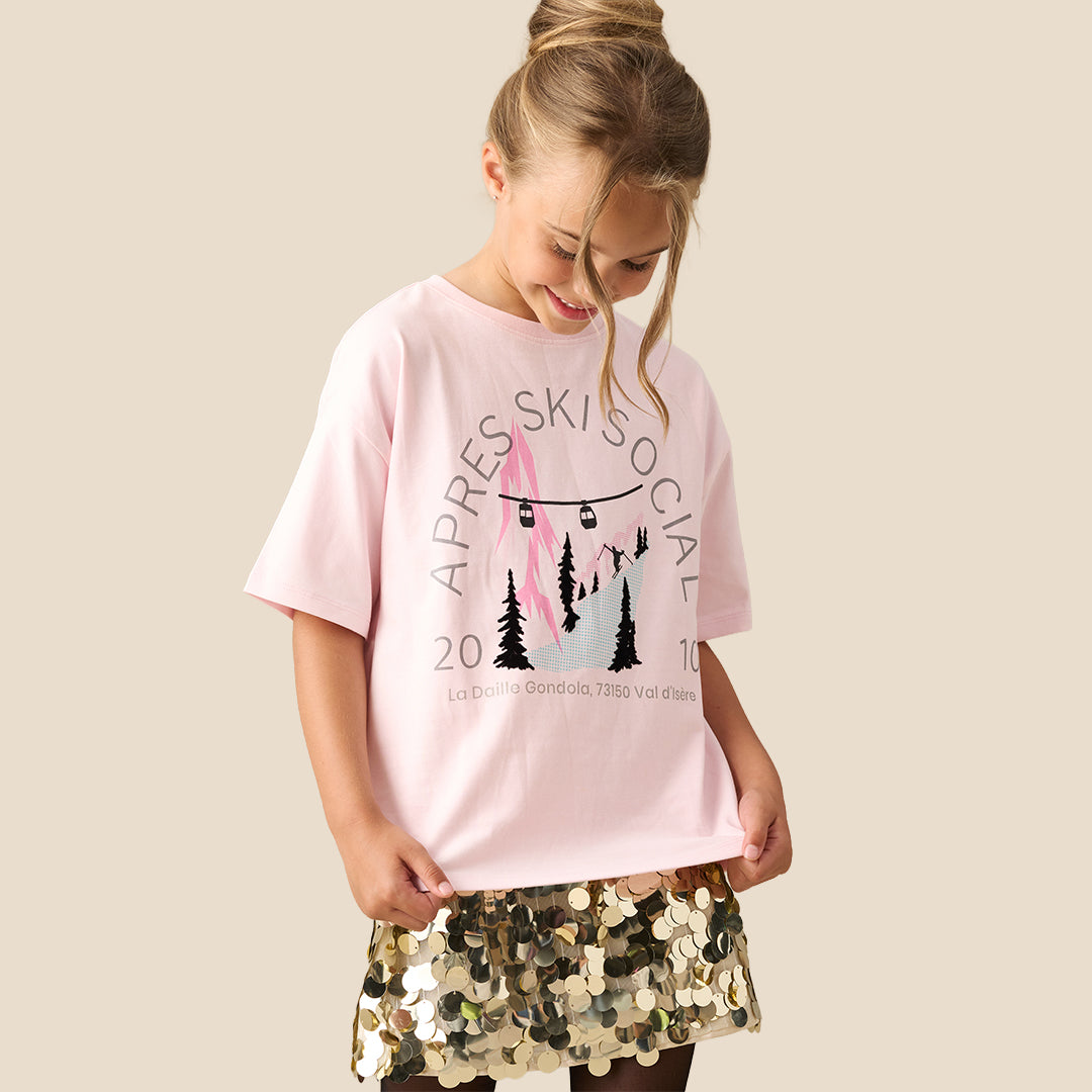 A young girl modeling a light pink graphic t-shirt with a black and pink ski design and text, paired with a gold sequined skirt, against a solid beige background.