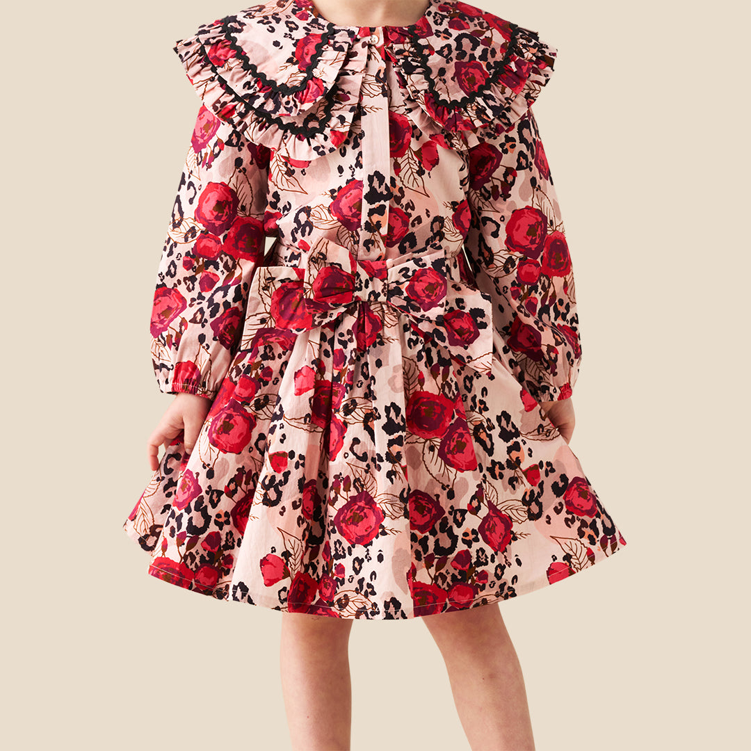 A young girl wearing a long-sleeve dress with a red rose and black leopard print pattern on a light pink background, photographed against a solid beige backdrop.