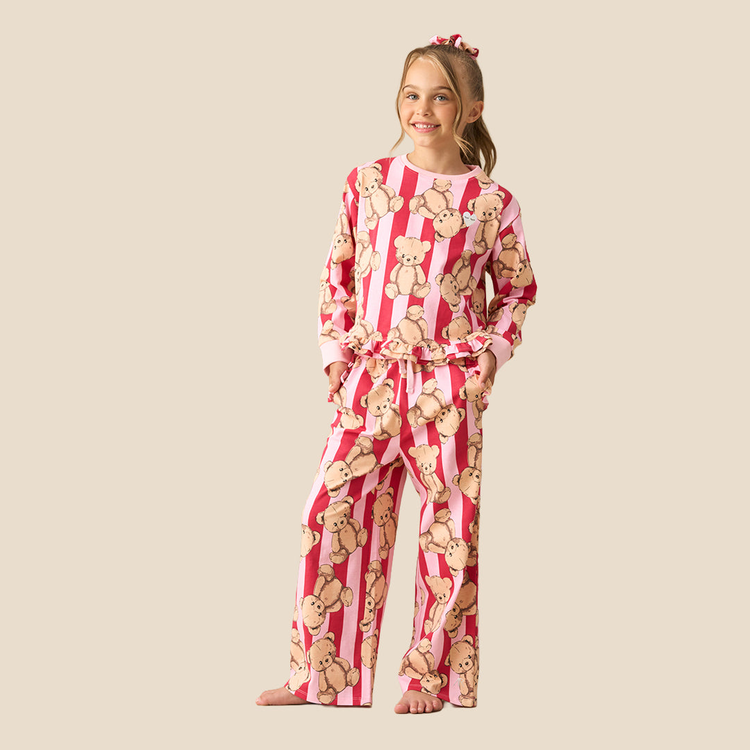 A young girl modeling a two-piece pajama set featuring a pink and white striped pattern with a repeating teddy bear print, set against a solid beige background.