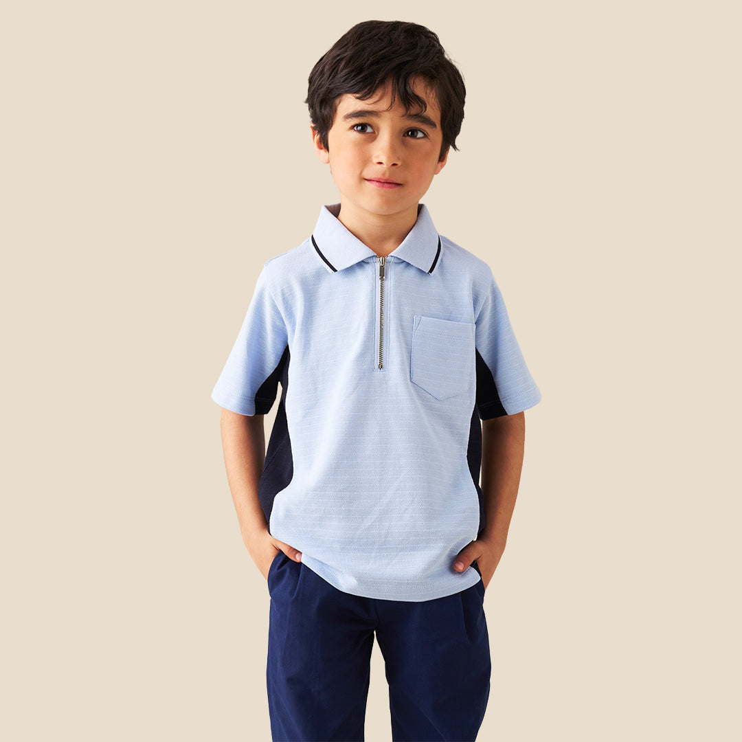 A young boy modeling a light blue polo shirt with dark navy side panels and a half-zip collar, paired with navy pants, against a solid beige background.