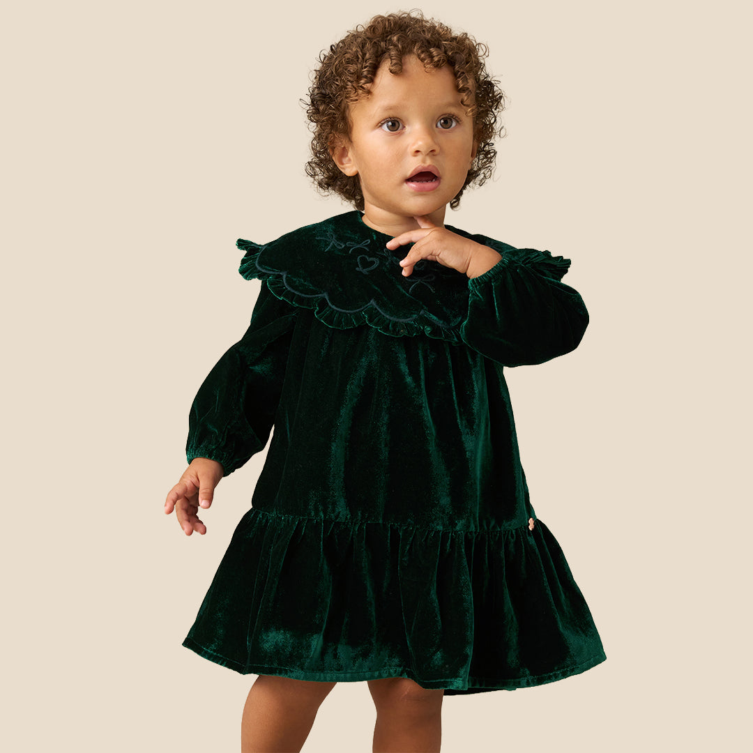 Two young children standing barefoot against a plain beige studio background, wearing matching dark green velvet dresses with wide ruffled shoulders and long sleeves.