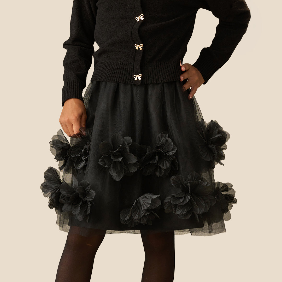 A model wearing a black cardigan with gold bow buttons and a black tulle skirt adorned with large black fabric flowers, set against a solid beige background.
