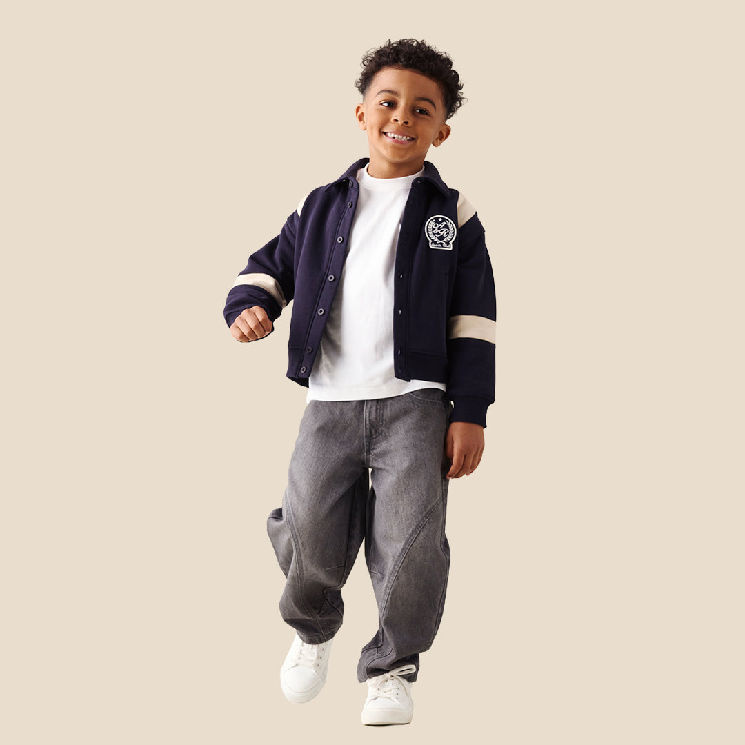 A child wearing baggy dark gray washed denim jeans with contrast stitching, styled with a navy and beige varsity jacket, white t-shirt, and white sneakers against a solid beige background.