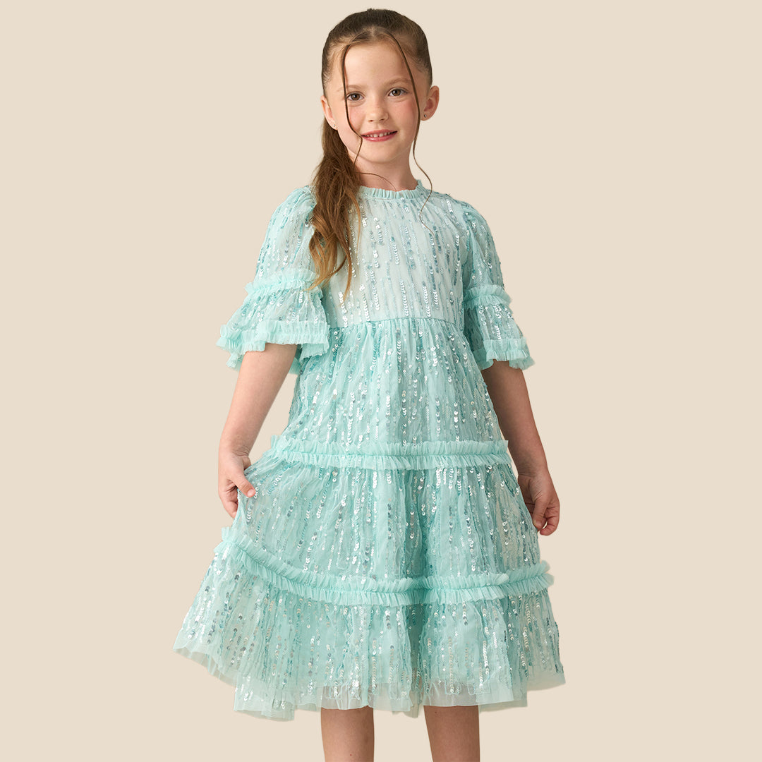 A young girl with a long brown ponytail wearing a light mint green sequined tiered dress, standing against a solid beige background.