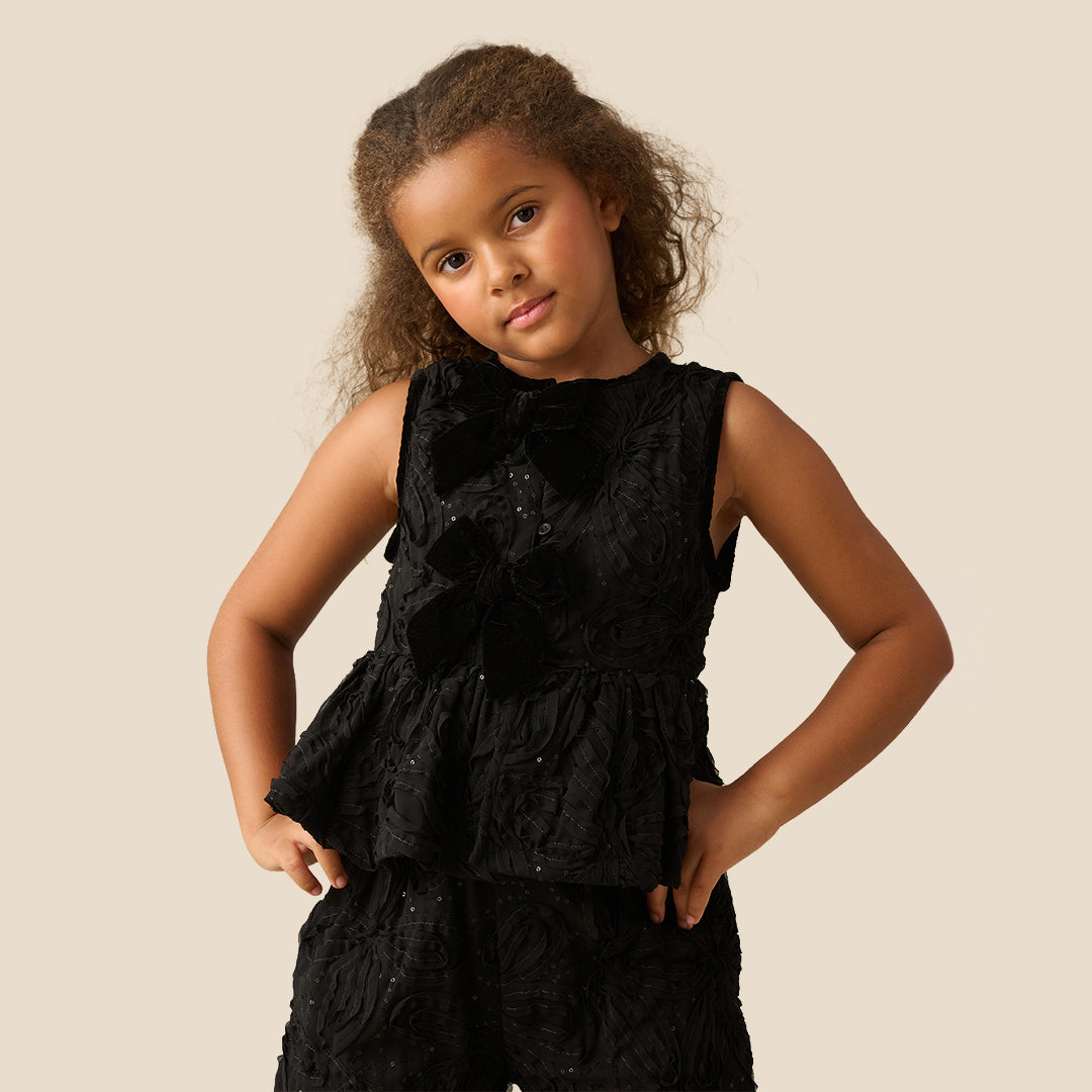 A young girl with curly brown hair wearing a sleeveless black textured dress with a ruffled peplum and bow detail, posing against a solid beige background.