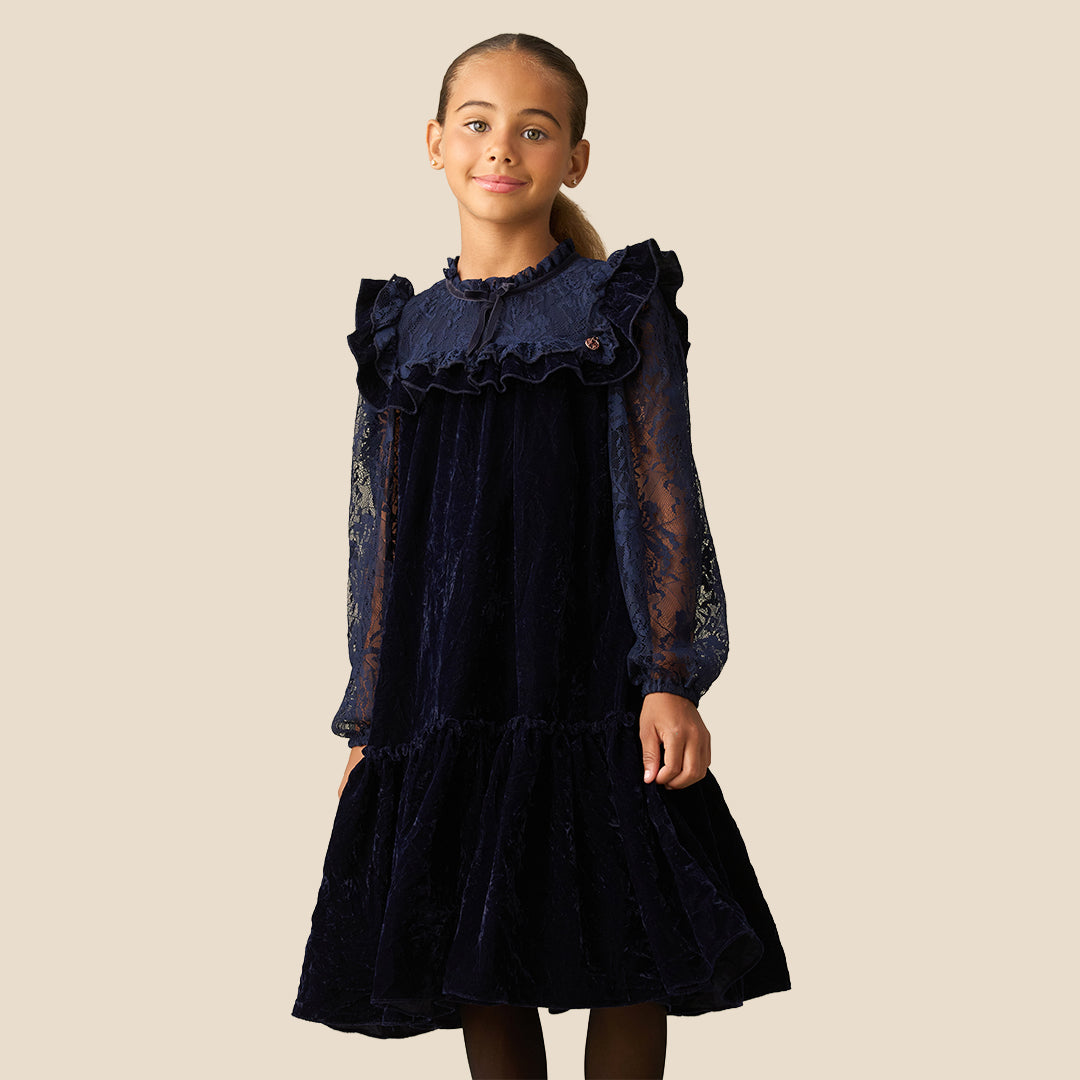 A young girl with her hair in a ponytail models a dark navy blue dress. The dress features a velvet-like texture, a ruffled collar, sheer lace long sleeves, and a tiered skirt, set against a solid beige background.