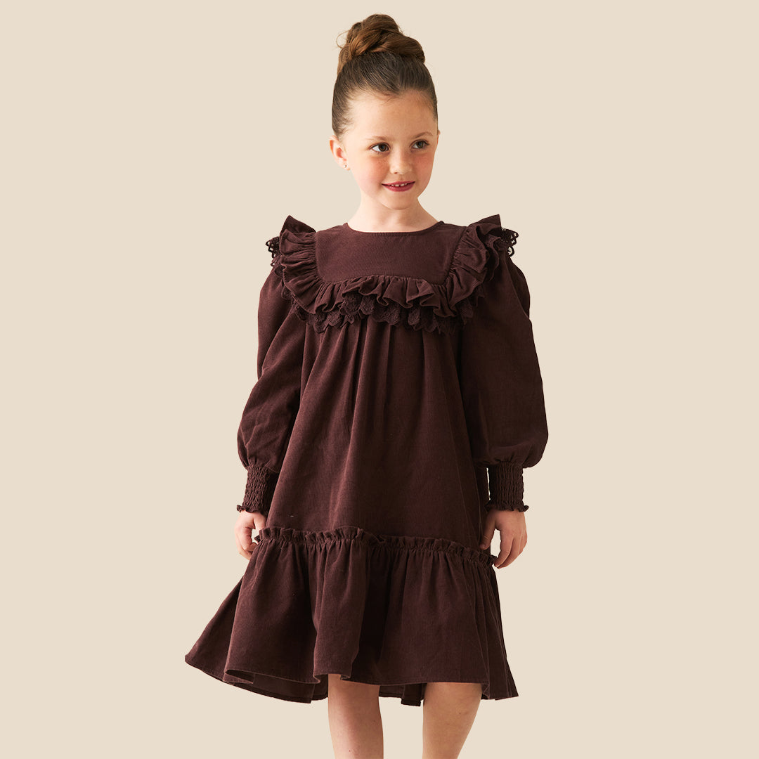 A young girl with her hair in a high bun models a dark brown long-sleeve dress with ruffled details, set against a solid light beige background.