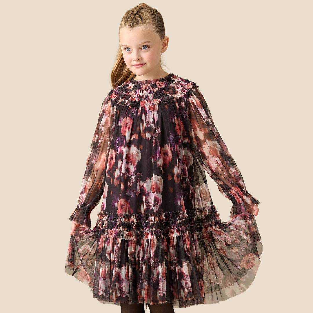 A young girl with her hair in a ponytail is wearing a long-sleeved, tiered tulle dress with a pink, red, and white floral pattern on a dark background, set against a solid beige studio backdrop.